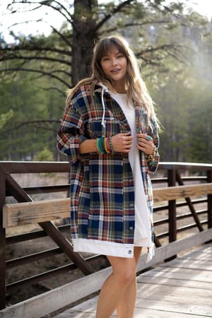 A woman stands on a wooden bridge outdoors in the Flannel Layers Hooded Jacket - Navy Multi over a white dress. She wears bracelets on both wrists, her hair is loose and wavy, and sunlight streams through the trees behind her.