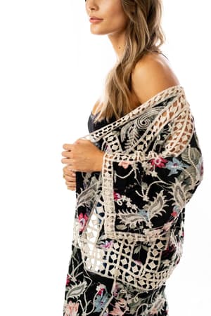 Woman with wavy light brown hair wears a black dress and the Let's Get Tropical Kimono—Black, featuring cream crochet and colorful floral embroidery, standing against a white background.