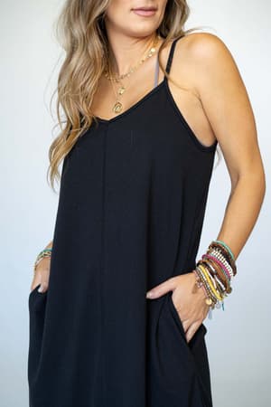 A woman wears The Nest Perfect Round Hem Bralette Maxi Dress in black, hands tucked into its pockets. She accessorizes with layered gold necklaces and colorful beaded bracelets, her long wavy hair styled simply against a plain light background.