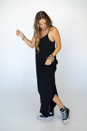 A woman with long wavy hair wears The Nest Perfect Round Hem Bralette Maxi Dress in black and black high-top sneakers. She lifts one side of her dress, showing colorful bracelets on her wrist, standing against a plain white background.