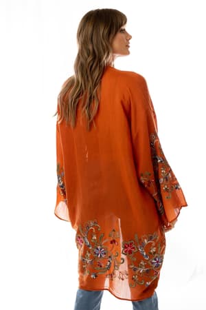 A woman with wavy, light brown hair is wearing the Fun In Florals Kimono - Rust, featuring colorful floral embroidery on the back and sleeves. The sheer rust fabric showcases the detailed design against a white background.