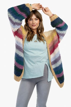 A woman in a pastel blue shirt, light gray jeans, and the Wrapped up in Color Knit Cardigan - Neutral—featuring a chunky, multicolored striped knit and relaxed fit—smiles with arms raised against a white background.