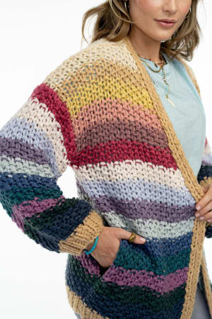 A woman wears the “Wrapped up in Color Knit Cardigan - Neutral,” a bold, chunky, hand-crocheted piece with colorful horizontal stripes and a loose fit. She pairs it with layered necklaces and has one hand in her pocket.