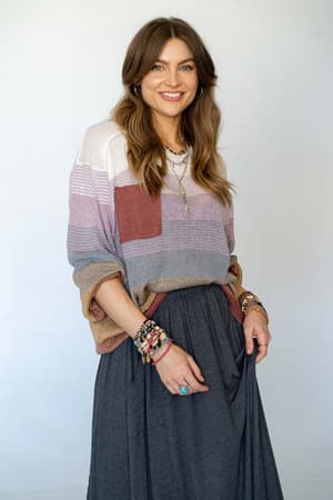A woman with long brown hair and layered necklaces smiles at the camera, wearing the Going Places Gradient Stripe Sweater in mauve and a long dark gray skirt, accessorized with multiple bracelets and rings. The background is plain light gray.