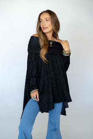 A woman with long brown hair wears The Nest So Versatile Flowy Knit Sweater in Charcoal and light blue jeans, accessorized with layered necklaces and colorful bracelets as she poses against a plain light background.