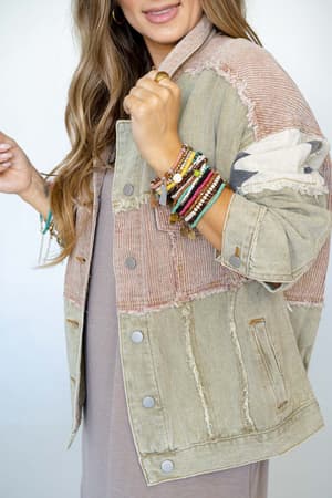 A woman with long wavy hair wears the Star Dust Contrast Corduroy Jacket - Multi over a taupe dress. She shows off her beaded bracelets and rings while holding the jacket’s collar and smiling softly.