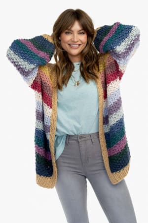 A woman smiles with her hands behind her head, wearing a light blue shirt, gray jeans, and the Wrapped up in Color Knit Cardigan - Neutral. The chunky knit features bold horizontal multicolored stripes, beige trim, and a loose fit against a plain white background.