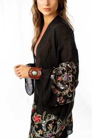 A woman models the Fun In Florals Kimono - Black, featuring vibrant embroidery on the sleeves and hem, against a white background. She has long, wavy hair and wears a statement bracelet.