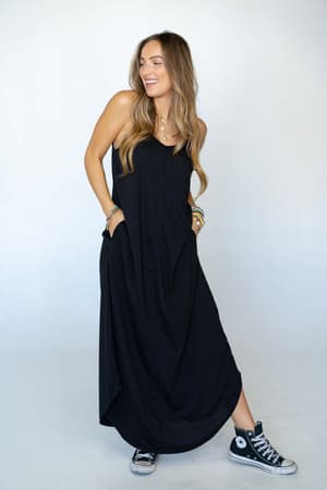 A woman with long, wavy hair smiles while wearing The Nest Perfect Round Hem Bralette Maxi Dress - Black and black Converse sneakers, standing against a light background.