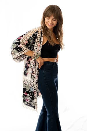 A woman with long brown hair smiles and looks down, wearing a black crop top, high-waisted flared jeans, and the Let's Get Tropical Kimono - Black with colorful floral print and wide sleeves against a white background.
