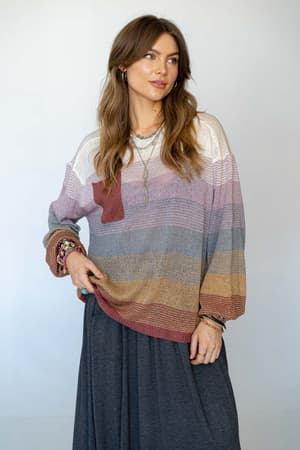 A woman with long, wavy brown hair wears the Going Places Gradient Stripe Sweater in Mauve and a long gray skirt, accessorized with bracelets and layered necklaces, standing against a plain light background.