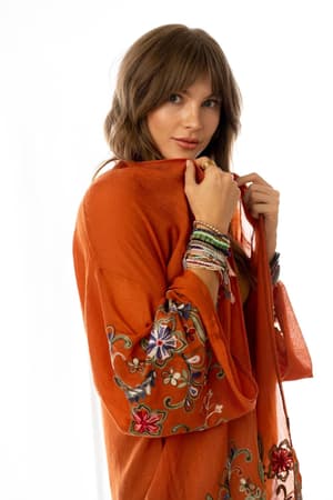 A woman with light brown hair wears the Fun In Florals Kimono - Rust, featuring embroidered floral patterns, and holds the sheer orange fabric near her chin. She accessorizes with colorful bracelets and faces the camera against a white background.