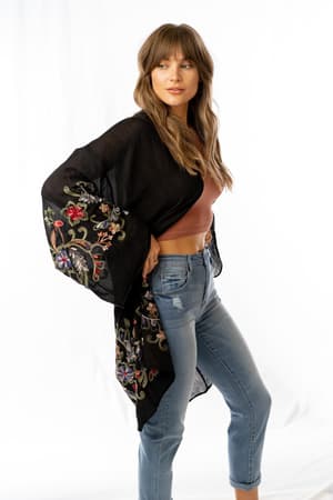 A woman with long brown hair models the Fun In Florals Kimono - Black over a mauve crop top and blue jeans. She poses against a white background, looking to the side with one hand on her hip.