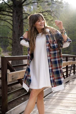 A smiling woman with long hair and bangs stands on a wooden bridge in a forest, wearing the Flannel Layers Hooded Jacket - Navy Multi over a white dress. She accessorizes with bracelets and looks off to the side with one arm raised.