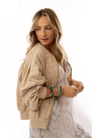 A woman with wavy blonde hair wears the Lorelei Crochet Jacket - Beige over a pale patterned dress, showing colorful beaded bracelets as she looks to the side against a white background.