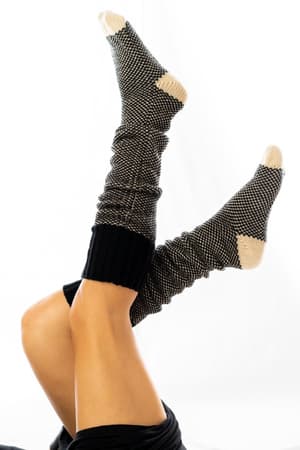 A person’s legs are raised in the air, wearing Cozy Cabin Lounge Socks - Black with black-and-white knit and beige toes and heels against a white background—perfect for a comfy, cabin-approved day.