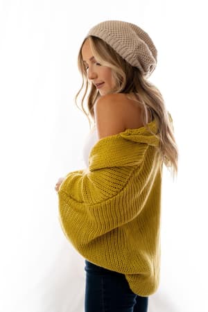 A woman with long, wavy blonde hair models the Cozy Hooded Cardigan in mustard, worn off one shoulder over a white top with a beige knit beanie, standing against a white background.