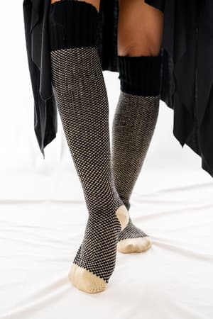 Someone stands on a white surface wearing Cozy Cabin Lounge Socks - Black, featuring tall black-and-white knit with beige toes and heels. Part of a black garment is also visible.
