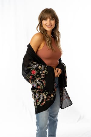 A woman with long light brown hair smiles, wearing a sleeveless brown top, light blue jeans, and the Fun In Florals Kimono - Black with colorful embroidery, standing against a plain white background.