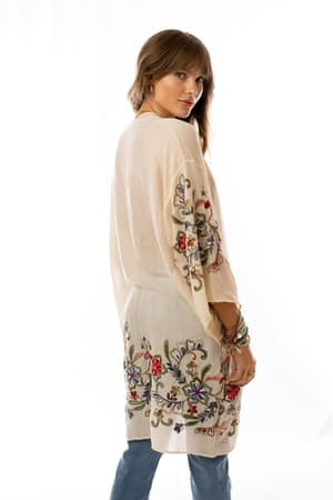 A woman stands slightly angled, wearing the Fun In Florals Kimono - Cream, a light, knee-length tunic with colorful floral embroidery on the sleeves and hem, paired with blue jeans against a plain white background.