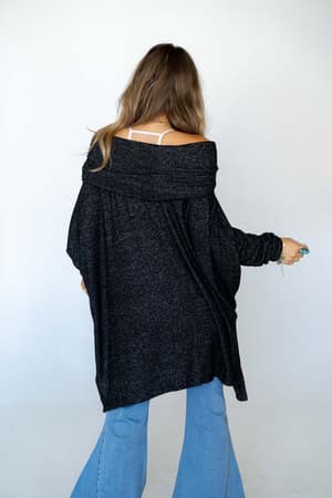 A woman with long hair stands facing away against a white background, wearing The Nest So Versatile Flowy Knit Sweater in charcoal off one shoulder, light blue flared jeans, and a visible white strap.