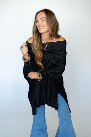 A woman with long brown hair smiles while wearing The Nest So Versatile Flowy Knit Sweater in Charcoal, paired with flared blue jeans and colorful accessories, standing against a plain light background.