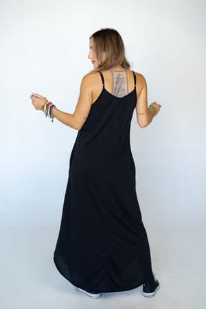 A woman with long hair stands facing away, wearing The Nest Perfect Round Hem Bralette Maxi Dress - Black and black sneakers. She has a large upper back tattoo and colorful bracelets on both arms, set against a plain white background.
