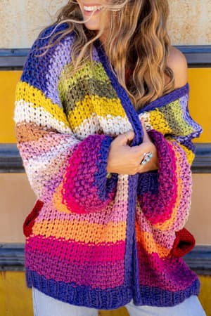 A woman with wavy hair smiles while wearing the Wrapped up in Color Knit Cardigan – Rainbow, a chunky, open front boho sweater with vibrant purple, pink, yellow, red, and blue stripes, standing near a yellow and beige wall.