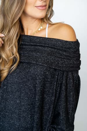 Wearing The Nest So Versatile Flowy Knit Sweater in Charcoal, a woman with wavy light brown hair shows off an off-the-shoulder look, a visible strap, and a layered gold necklace for a soft, boho-inspired style.