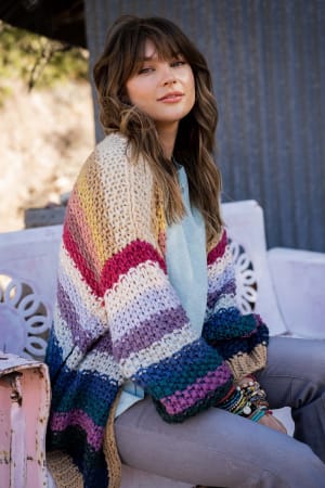 A woman with long brown hair sits on a white bench outdoors, wearing the Wrapped up in Color Knit Cardigan - Neutral over a light blue top and gray pants. She accessorizes with bracelets and gazes at the camera with a relaxed expression.