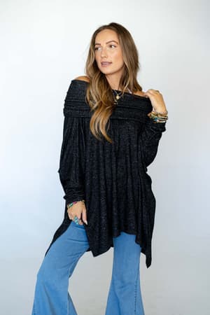 A woman with long brown hair wears The Nest So Versatile Flowy Knit Sweater in charcoal and light blue jeans, accessorized with boho-style bracelets and rings, standing against a plain light background.