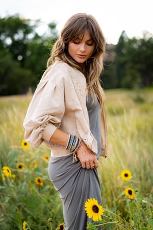 A woman with wavy brown hair stands in a field of yellow wildflowers, wearing the Lorelei Crochet Jacket - Beige over her grey dress and colorful bracelets. She looks down serenely, surrounded by tall grass and sunflowers.