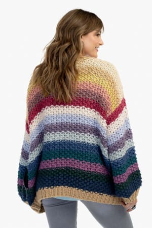A woman with long brown hair, seen from behind, wears the Wrapped up in Color Knit Cardigan - Neutral, featuring bold colorful stripes and beige trim. The loose-fit cardigan is styled with grey pants as she smiles slightly to the side.