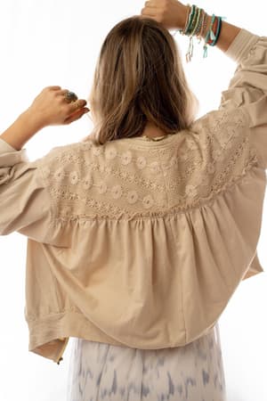 A woman with long hair, seen from behind, lifts her arms. She wears the Lorelei Crochet Jacket - Beige over a patterned dress and several colorful bracelets on her right wrist. The white background is bright.
