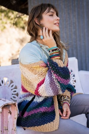 A woman sits on a white bench outdoors, smiling slightly as sunlight highlights her face. She wears the Wrapped up in Color Knit Cardigan - Neutral over a light blue top, paired with beaded bracelets and rings.