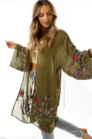 A woman with long, wavy blonde hair stands barefoot against a white background wearing the Fun In Florals Kimono - Olive, featuring colorful embroidery over a white top and light-wash ripped jeans.