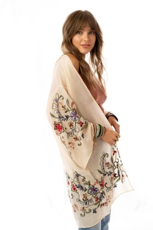 A woman with long, wavy hair wears the Fun In Florals Kimono - Cream, featuring sheer fabric and colorful floral embroidery over a pink top and blue jeans. She stands against a white background, smiling slightly with bracelets on her wrist.