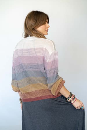 A woman with light brown hair stands to the side, wearing the Going Places Gradient Stripe Sweater in mauve and a long gray skirt. She holds her skirt and has colorful bracelets on her wrists. The background is plain and light.