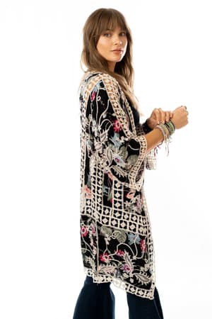 Woman with light brown hair wears the Let's Get Tropical Kimono - Black, featuring a floral print, colorful embroidery, and cream crochet trim. She stands against a white background, looking at the camera with bracelets on her wrist.