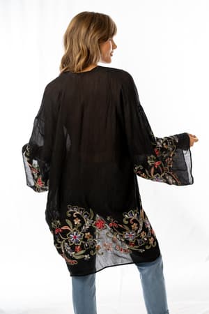 A woman with shoulder-length hair is seen from the back in the Fun In Florals Kimono - Black, featuring wide sleeves and colorful embroidery on sheer fabric, styled with light blue jeans against a white background.