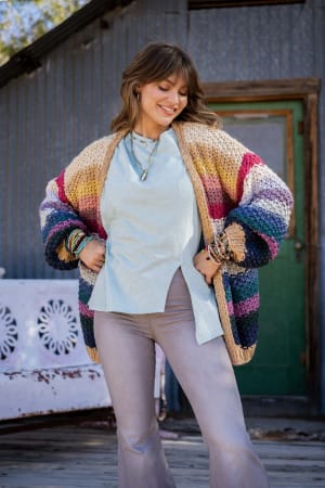 A woman stands outdoors smiling, wearing a light blue shirt, flared pants, and the Wrapped up in Color Knit Cardigan - Neutral, a bold, loose-fit striped knit in earth tones. She has long hair with bangs and wears bracelets and necklaces.