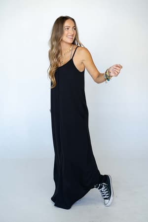 A woman with long wavy hair wears The Nest Perfect Round Hem Bralette Maxi Dress in black, styled with black high-top sneakers. She smiles and looks to the side while appearing mid-step against a plain white background.