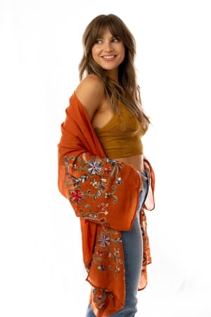 A smiling woman in profile wears a mustard yellow crop top, blue jeans, and the Fun In Florals Kimono - Rust draped over her shoulders. She has long, wavy brown hair and poses against a plain white background.