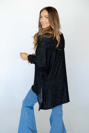 A woman with long, light brown hair smiles over her shoulder in The Nest So Versatile Flowy Knit Sweater - Charcoal and light blue flared jeans, creating a relaxed boho style against a plain white background.