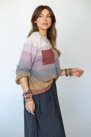 A woman with long brown hair wears the Going Places Gradient Stripe Sweater in mauve, featuring earth-tone stripes and a reddish pocket, paired with a long gray skirt and bracelets, standing sideways and looking off to the side.