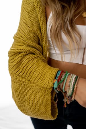 A woman styles the Cozy Hooded Cardigan - Mustard over a white crop top, accessorized with colorful beaded bracelets. The photo highlights her cozy look and the cardigan’s chunky knit, with her long wavy hair partially in view.