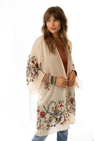 A woman with wavy light brown hair models the Fun In Florals Kimono - Cream, a sheer floral-embroidered layer styled over a top and jeans. She accessorizes with colorful bracelets and rings against a white background.
