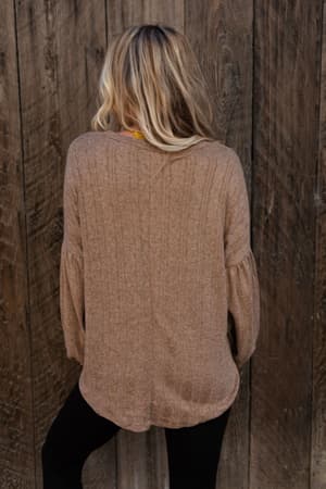A person with long, wavy blonde hair stands with their back to the camera, wearing The Nest Sweet Heart Balloon Sleeve Knit Sweater in Mocha and black pants while facing a wooden wall.