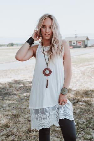 A woman with long blonde hair poses outdoors, wearing the Prima Lace Hem Slip in Ivory over black leggings and accessorized with layered necklaces, a bracelet, and a large wrist cuff. She touches her hair with one hand.