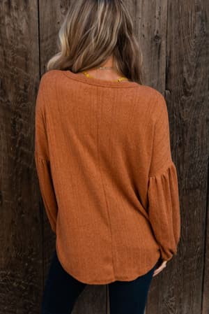 A person with long, wavy blonde hair stands by a rustic wooden wall, wearing The Nest Sweet Heart Balloon Sleeve Knit Sweater in burnt orange and dark jeans.